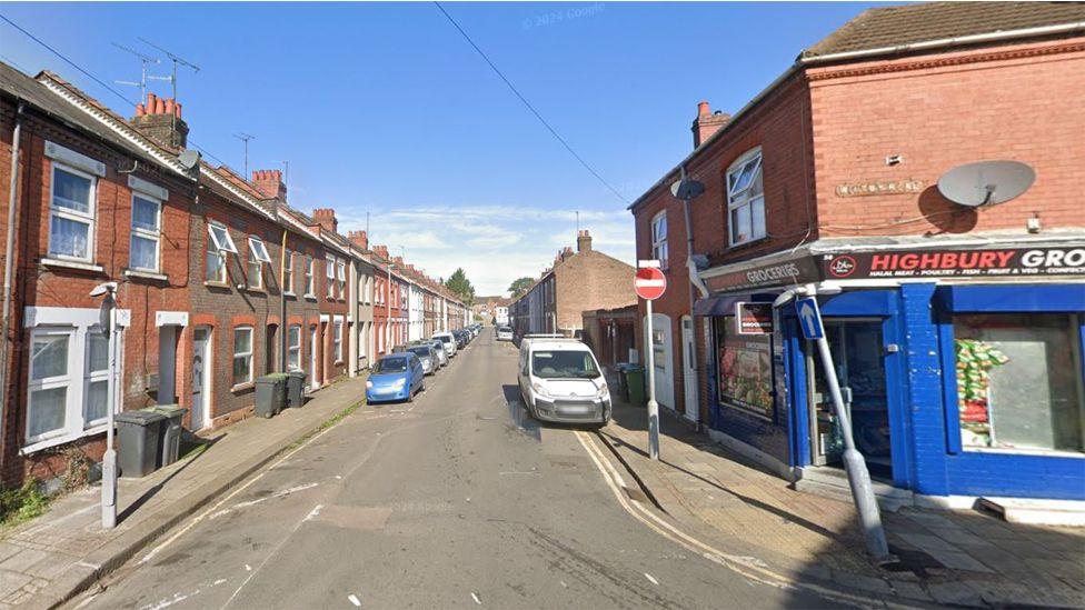 A terraced street with a long row of small brown and red brick terraced houses on the left side, tapering into the distance. On the right side is a blue corner shop titled Highbury Grocery. Cars line the left side of the street along with some wonky one-way street signs.