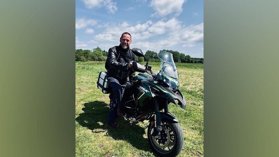 Ken Pottle is sitting on a large motorcycle in a field. He is wearing a black leather jackets and there are trees in the background