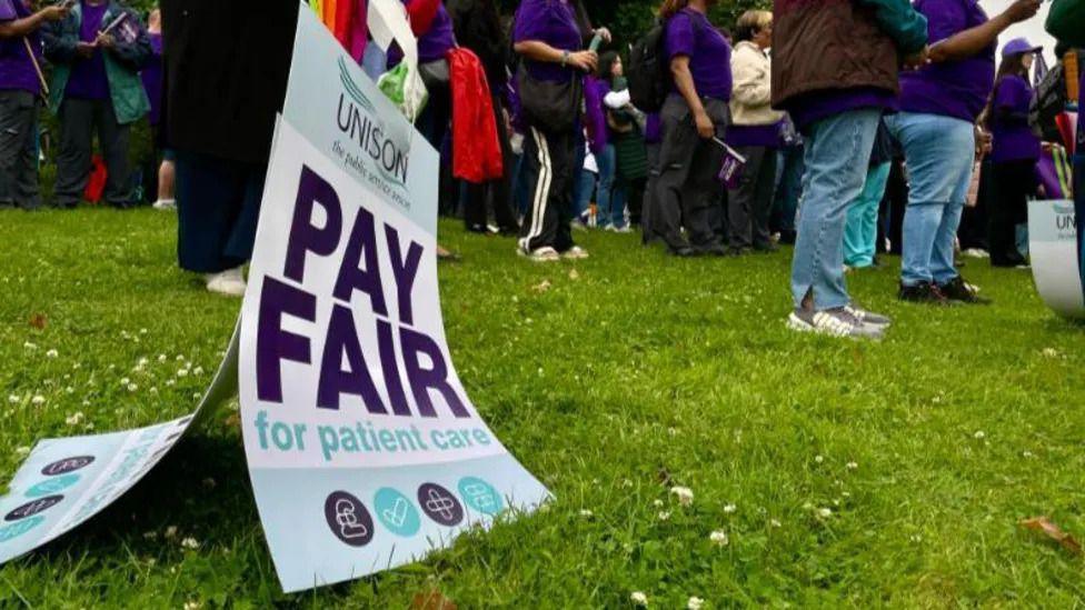 A sign saying "Pay fair for patient care" by workers feet on a picket line