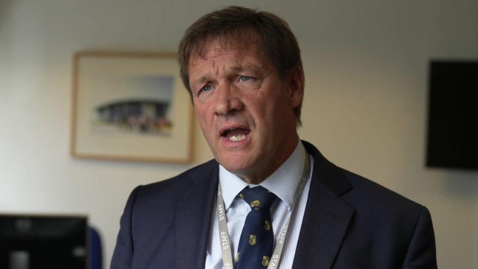 Martin Tucker, a middle-aged man with short brown hair, is dressed in a navy blue suit, white shirt, and a tie featuring crests. 