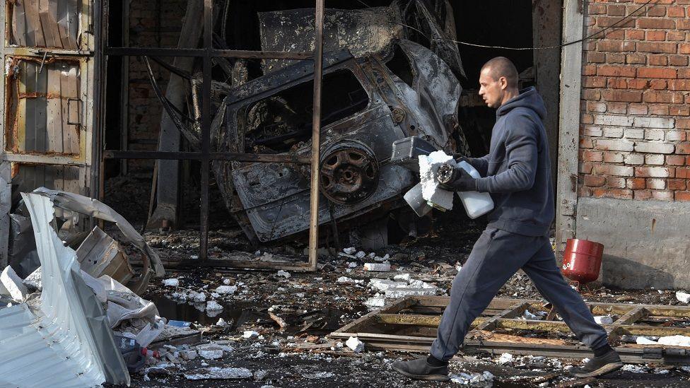 A man walks past a car repair workshop which was damaged by a Russian drone strike. The floor is covered in debris and there's a burnt out car in the background