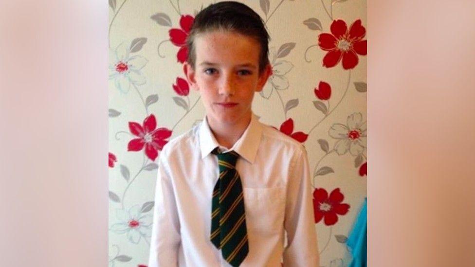 Tyler Kerry as a child stood after a red and silver floral patterned wall papered wall. He is wearing a white shirt and green and yellow striped tie.