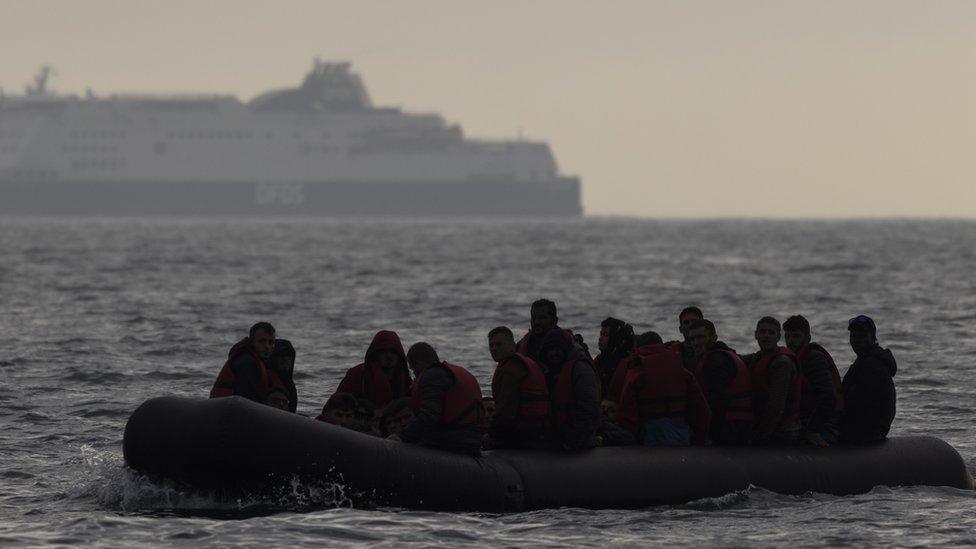 A group of about 20 people in an inflatable dinghy on the English Channel. It is dark and only their silhouttes can be seen. In the background a large ferry can be seen