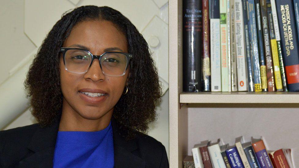 Dr Malisa Neptune-Figaro, wearing glasses and a blue shirt and black jacket, stands next to a book shelf at the University of the West Indies 