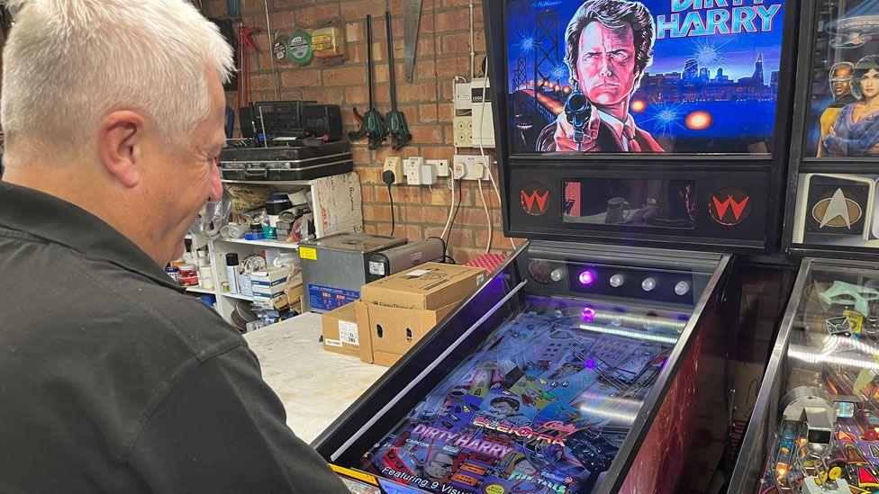 A side view of Mark Squires with short white hair, wearing a grey polo shirt, in front of a Dirty Harry pinball machine