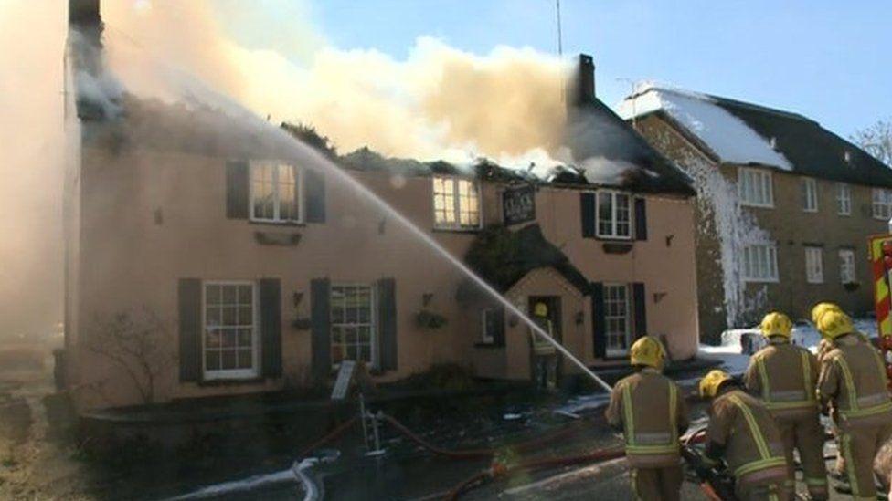 Firefighters spraying water onto the smoking thathed roof of the pink pub