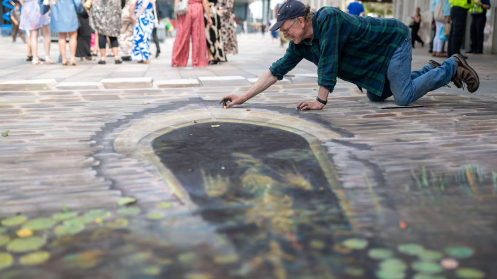 Artwork showing a water gate can be seen on the ground drawn in chalk. A man is drawing it as he kneels on the ground. He is wearing a green and blue shirt with a baseball cap on. There are people stood in groups in the background.