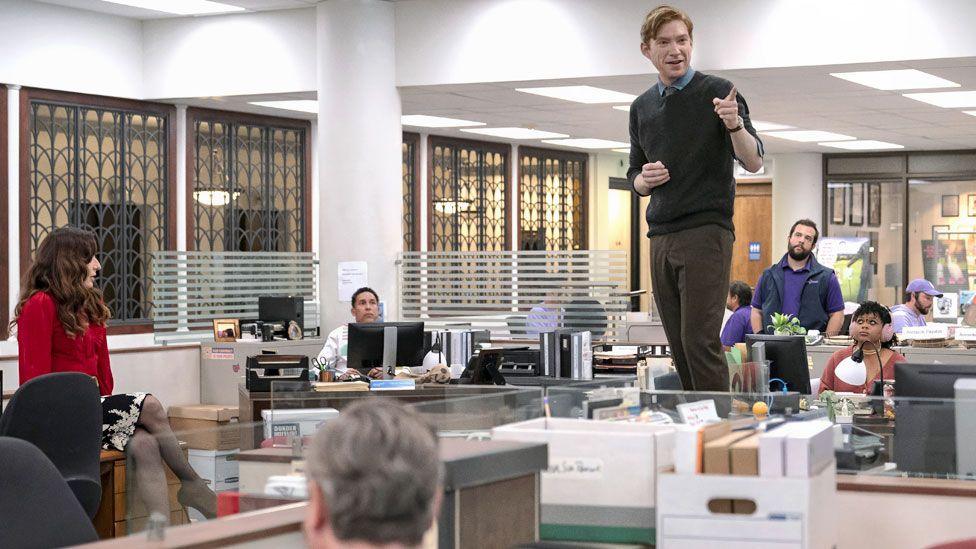 Domhnall Gleeson standing on a table in an office in character as Ned Sampson