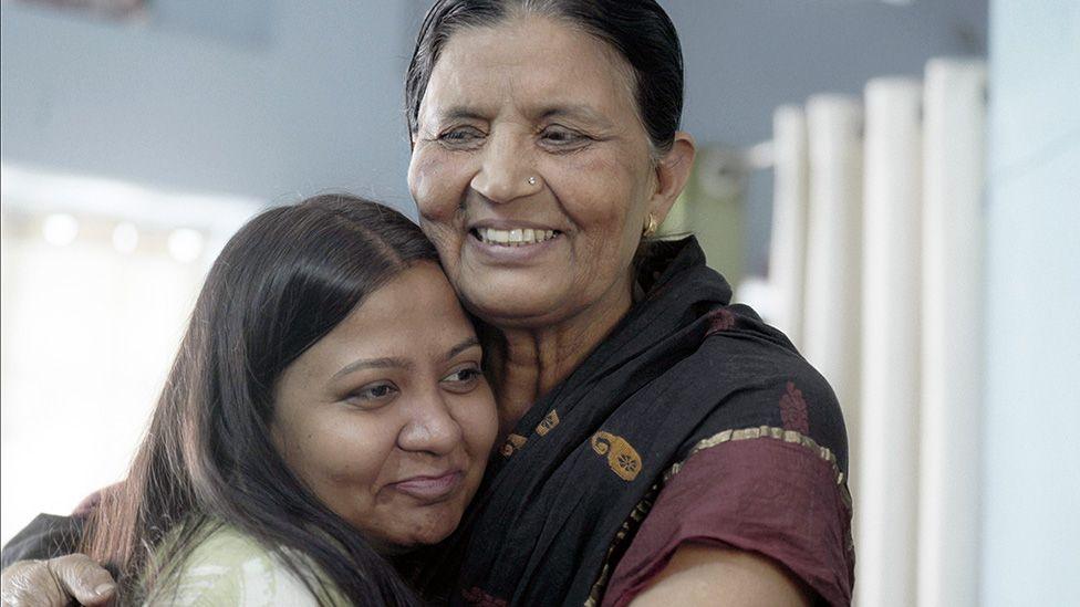 Anila - dressed in a black and maroon sari - and Monica hugging and smiling  