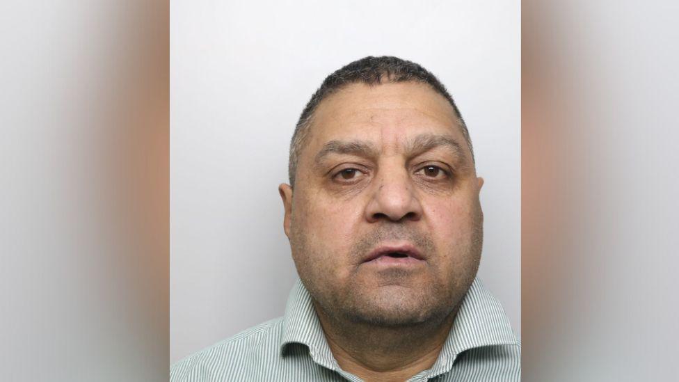 A police mugshot of a middle-aged man with a plump face and short grey hair. He is wearing an open-necked collar shirt.