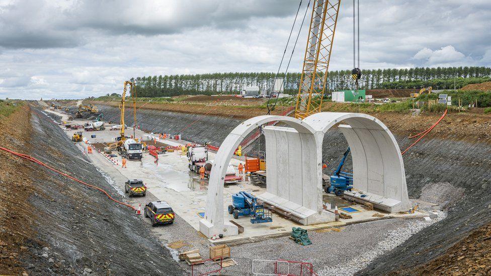 A huge concrete structure resembles an M shape. It is cut into a manmade valley, with lots of trucks and other vehicles parked. In the background on higher ground is a line of trees. It is cloudy.