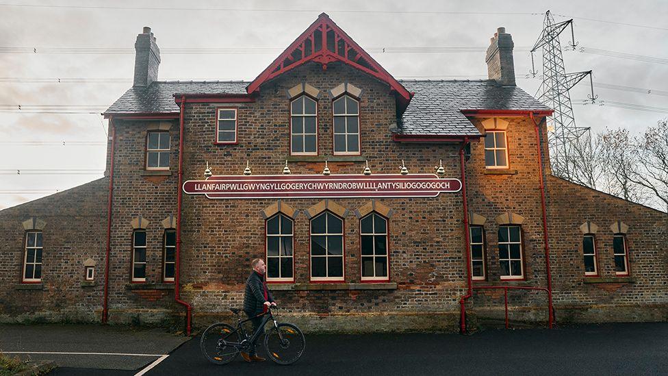 Cynghorydd Llanfairpwll Dyfed Wyn Jones yn cerdded heibio gorsaf drên adnabyddus y pentref