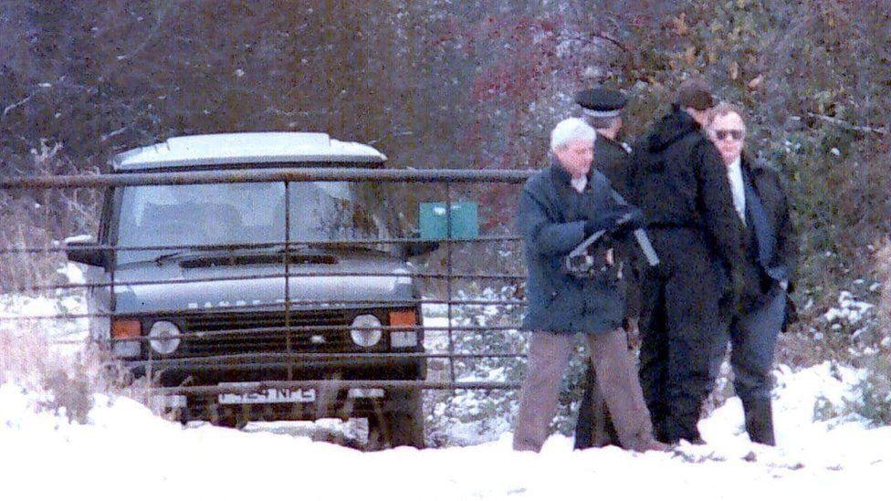 Uniformed police and detectives are standing in snow on farmland. Behind them is a metal gate and a Range Rover.