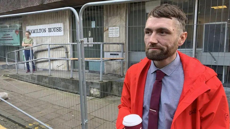 Wayne Lammond with a coffee in his hand standing outside a building in Chester he occupied as part of a homeless squat. He's wearing a red waterproof coat, purple shirt and maroon tie with a tie clip. He has short brown hair and a neat beard.