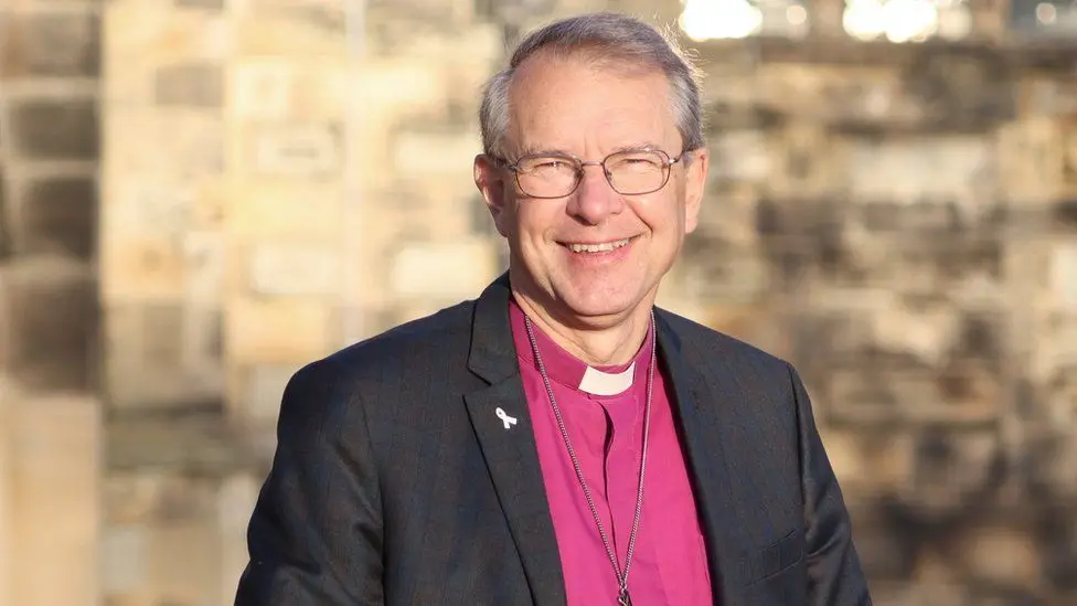 The Right Reverend Paul Butler wearing a reverend's collar and black blazer. He is smiling and wears glasses. He has a purple top on and a chain. 