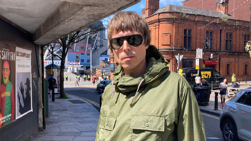A picture of Glenn looking at the camera with a stern expression. He is wearing a khaki green parka jacket, black sunglasses and has short brown hair. He is standing in Cardiff town centre. Behind him is a red brick building and the Principality Stadium.