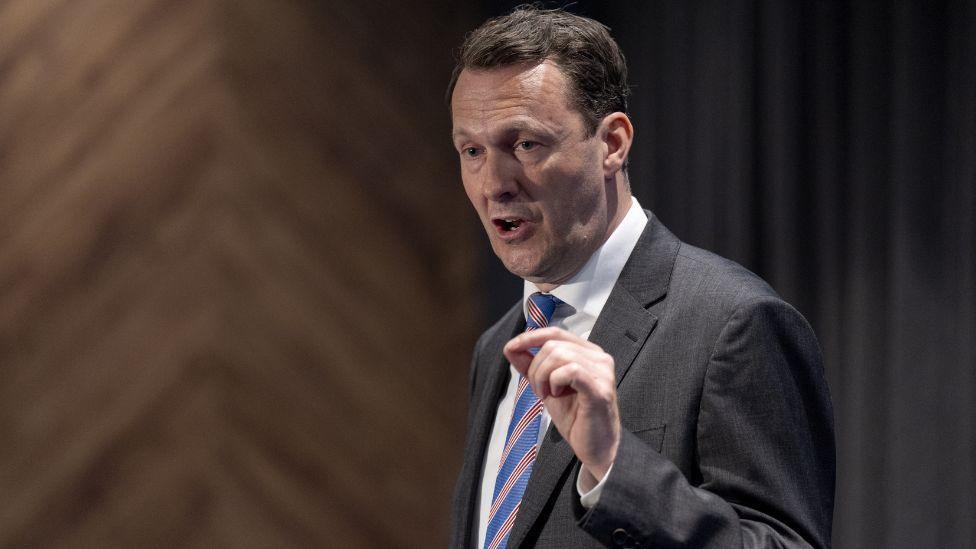 Russell Findlay, a man with dark hair, speaks with his left hand raised. He is wearing a dark suit, white shirt and blue and red stripy tie. There are curtains and wood panelling behind him. 