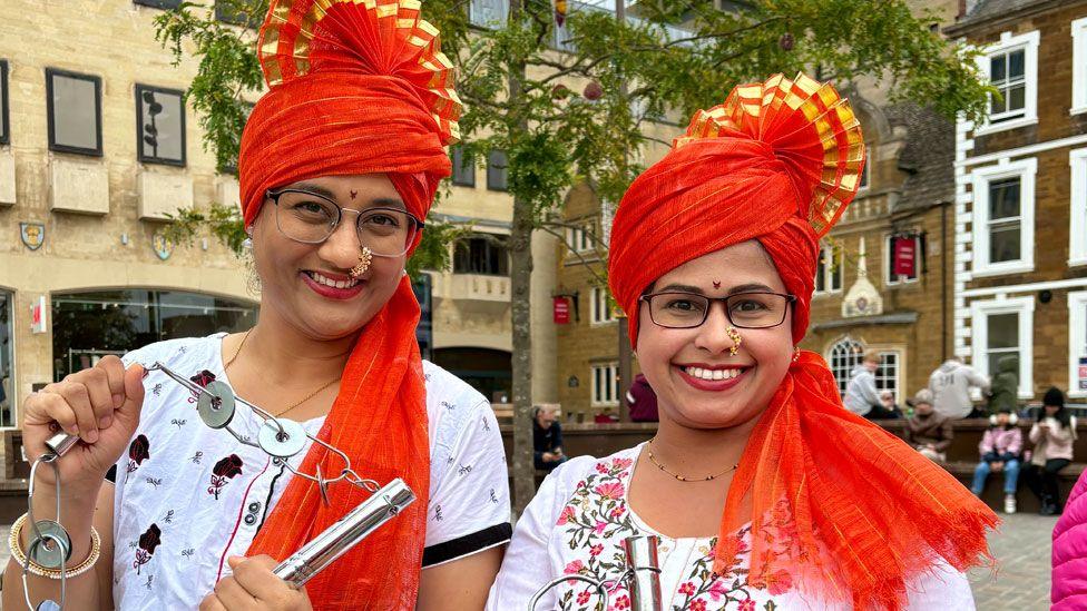 Two smiling women, standing in Northampton's Market Square. They are wearing orange headdresses which have gold tipped pleats in a comb on the top and white embroidered tops. Both are wearing glasses.