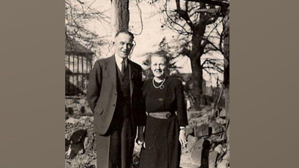 A grainy black and white photo shows Gordon and Ruth Wimbush standing together in what looks like a park. There are trees, rocks and part of a pavilion behind them, He is wearing a dark suit and she is wearing a dark dress. The couple look to be in their 50s or 60s.