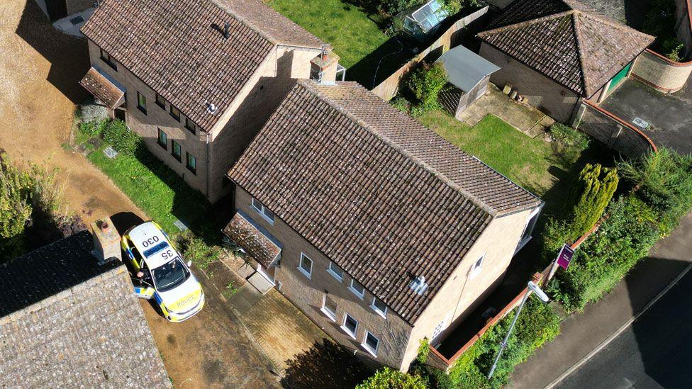 Aerial shot  showing a police car on the right and the roof of a detached two-storey house and its garden beyond, John Amner Close, Ely