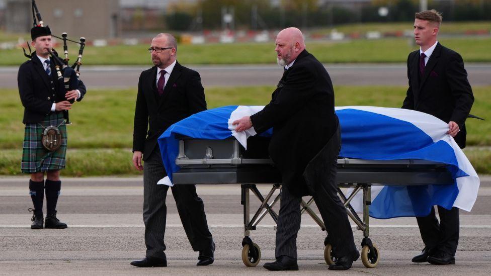 Alex Salmond's coffin draped in a saltire at Aberdeen airport, with a bagpiper in the background