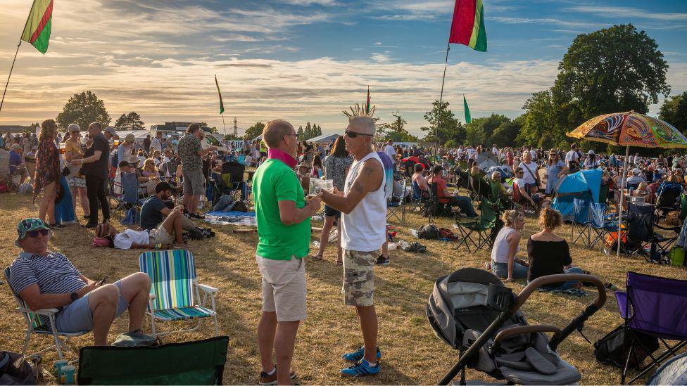 A large mass of people stood and sitting in a field on the site of GuilFest. There are hundreds of people in a large crowd. 