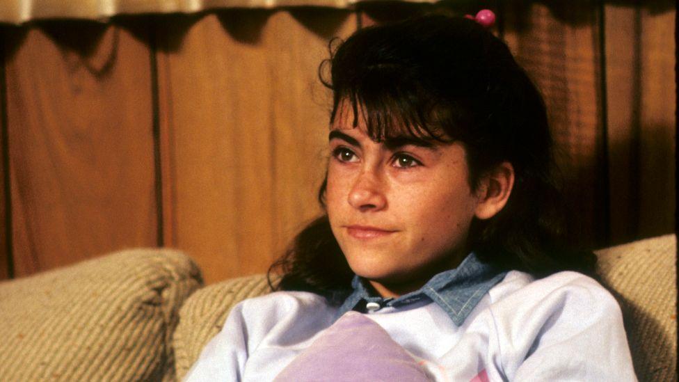 Roxana Zal, pictured in 1984's Something About Amelia, sitting on a sofa, wearing a white jumper with her black hair tied up