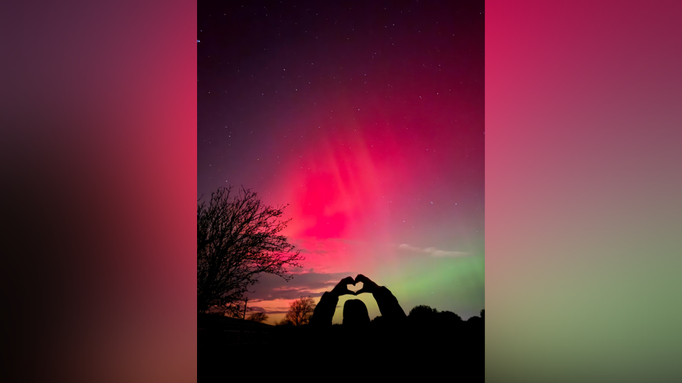 A silhouette of a person holding up a love heart directly towards a backdrop of the Northern Lights in bright red, bright green and darker orange and purple tones