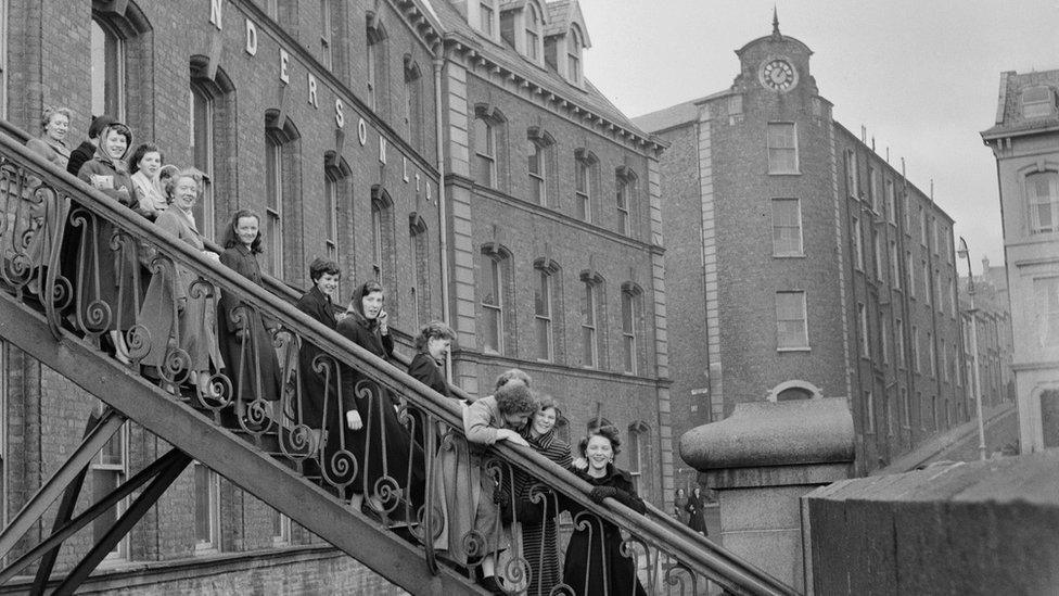 Workers leaving Tillie and Henderson's shirt factory in November 1955 stop on the factory steps for a photo. There are around 15 women, dressed on overcoats. The old factory building is in the background