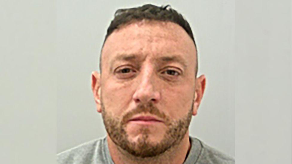 Jake Balmforth is looking straight at the camera in this police mugshot. He has short brown hair and stubble and is wearing a grey top.
