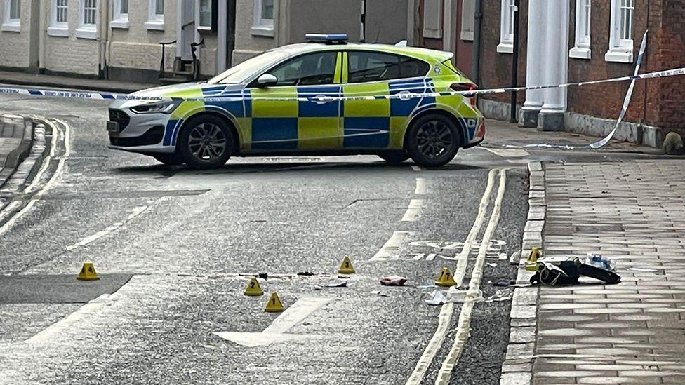 A view of the scene in Beverley with a police car and cordon tape with evidence markers on the road and pavement