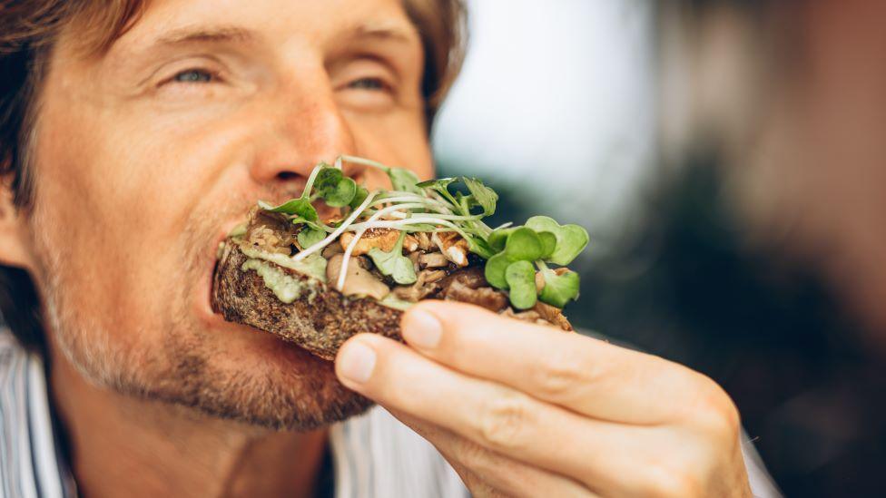 A close-up of a man eating an open sandwich with mushrooms and leaves