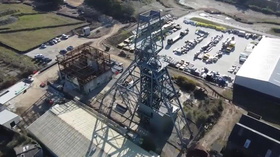 A pit shaft metal gear above the surface with a mass of metal girders and a car park behind along with a number of industrial buildings.