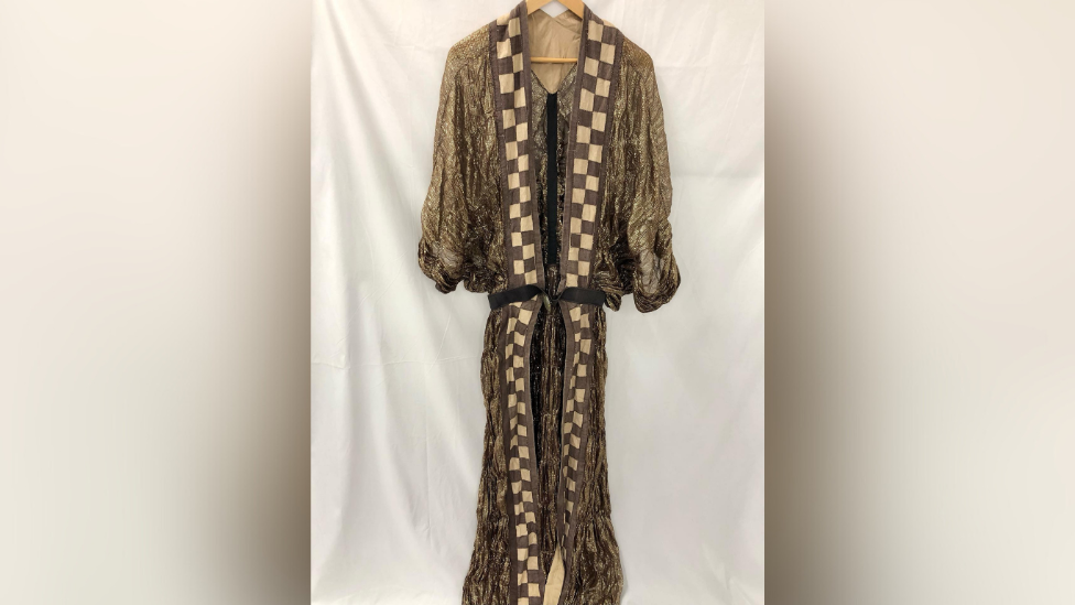 A long theatre garment is hung on a hanger in front of a white cloth. It has chequered brown and white squares around the neckline and front of the clothing. The rest of it is glittery. 