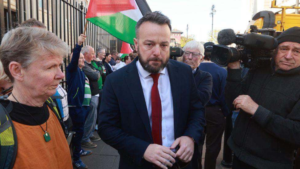 Colum Eastwood pictured outside court, he is in a navy suit with a red tie and white shirt. He has short, dark hair and a brown beard. There are numerous cameramen in dark clothing, holding cameras beside him. There are also numerous supporters, waving black red white and green Palestine flags. Directly to Colum's right is a short haired woman wearing an orange top.