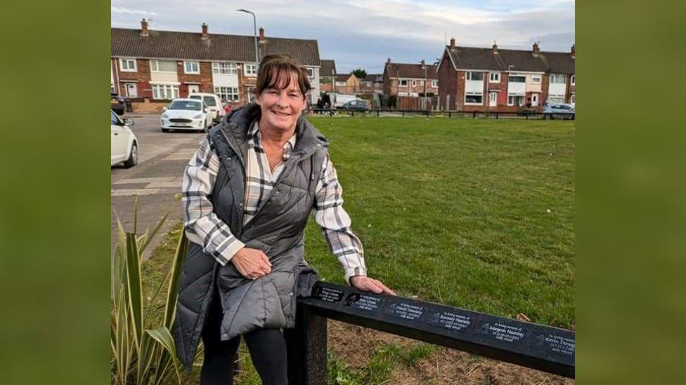 Ann Hanratty wearing a white check shirt and a jacket posing in a park. She is next to a black fence which has the plaques bearing the names of local people on it. 