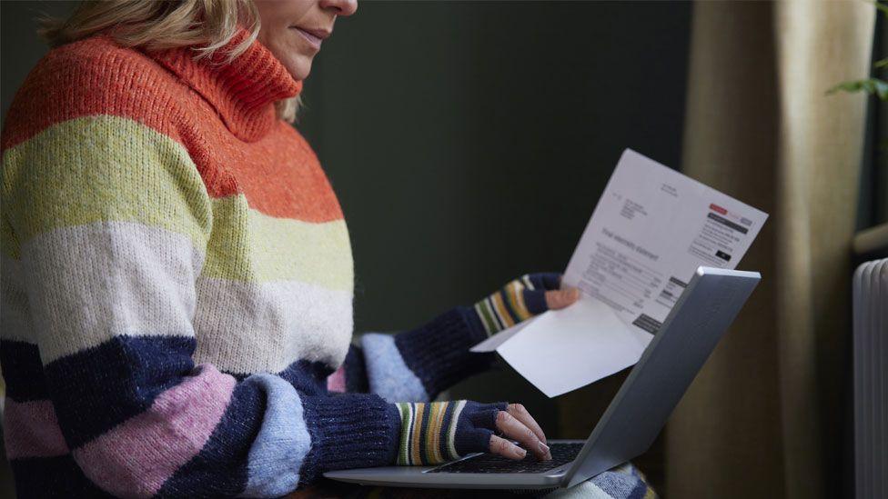 A woman sits in her home wearing a stripey jumper and gloves whilst reading a bill with her laptop on her lap