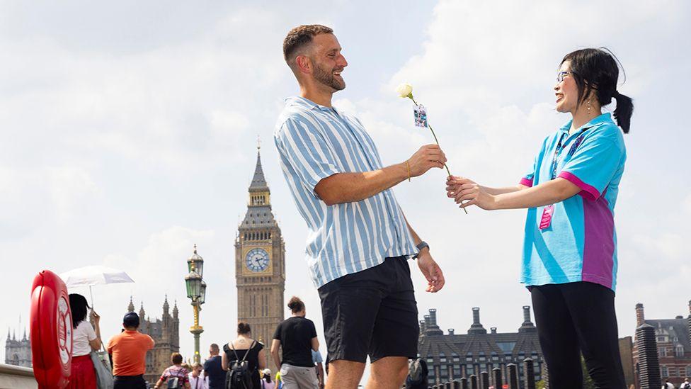 A Hull volunteer in a blue shirt handing a white rose to a man on Westminster Bridge with Big Ben in the background