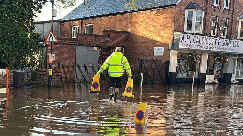 Man in high-vis jacket and wellies puts out cones in flooded street