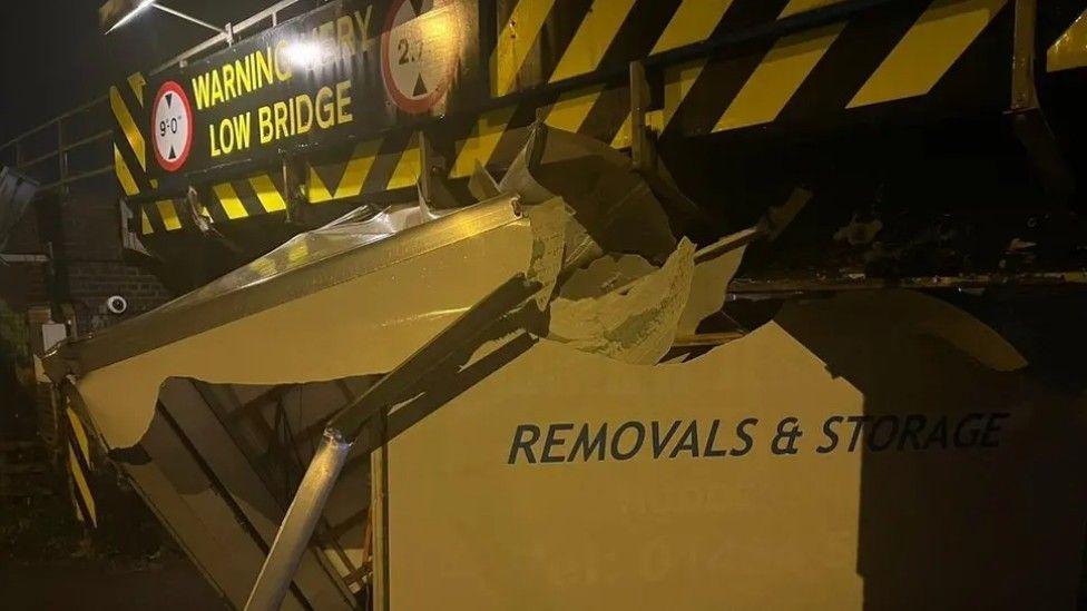 A partially destroyed van is wedged under a bridge. The bridge has a large warning sign on it informing drivers that it is "very low"