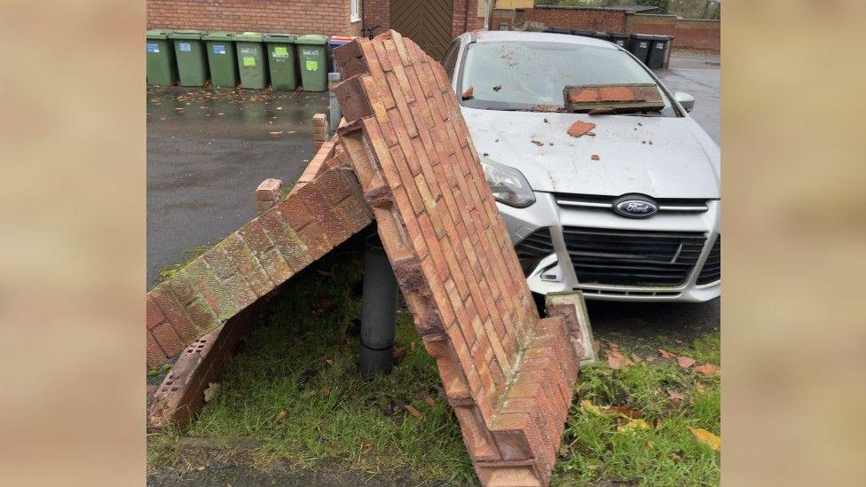 A brick wall  which has fallen over. Brick debris is on the windscreen of a silver Ford car.