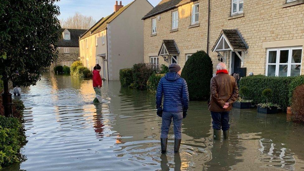 Three people stand in flood water outside a row of brick houses. The water reaches above their ankles and they are all wearing welly boots. 
