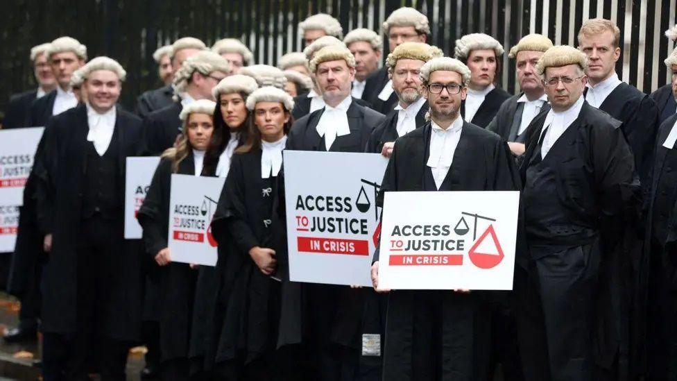 A number of barristers, both male and female, standing in black robes and white wigs, a number of them holding placards saying access top justice in crisis