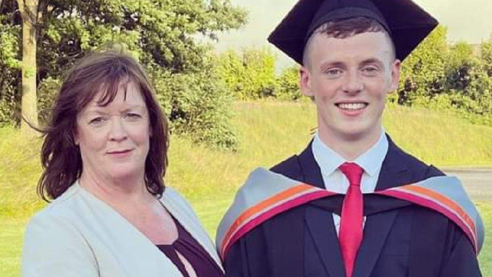 Lesley Mease and her son, Wilson Shepherd, stand together against the backdrop of a park. Lesley is wearing a suit and Wilson is wearing graduation robes. He has short dark hair.