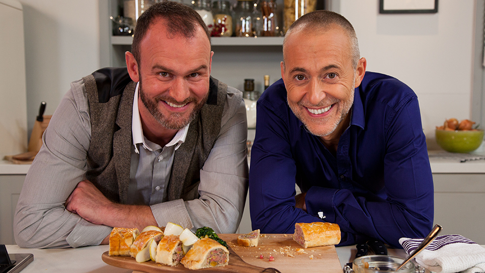 Glynn Purnell and Michel Roux Junior leaning on a worktop, with food in front of them  on  chopping board