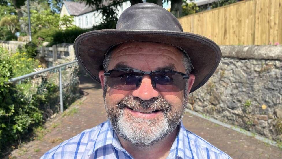 A man with a blue checked shirt, wearing a white brimmed hat and sunglasses. He is wearing sunglasses