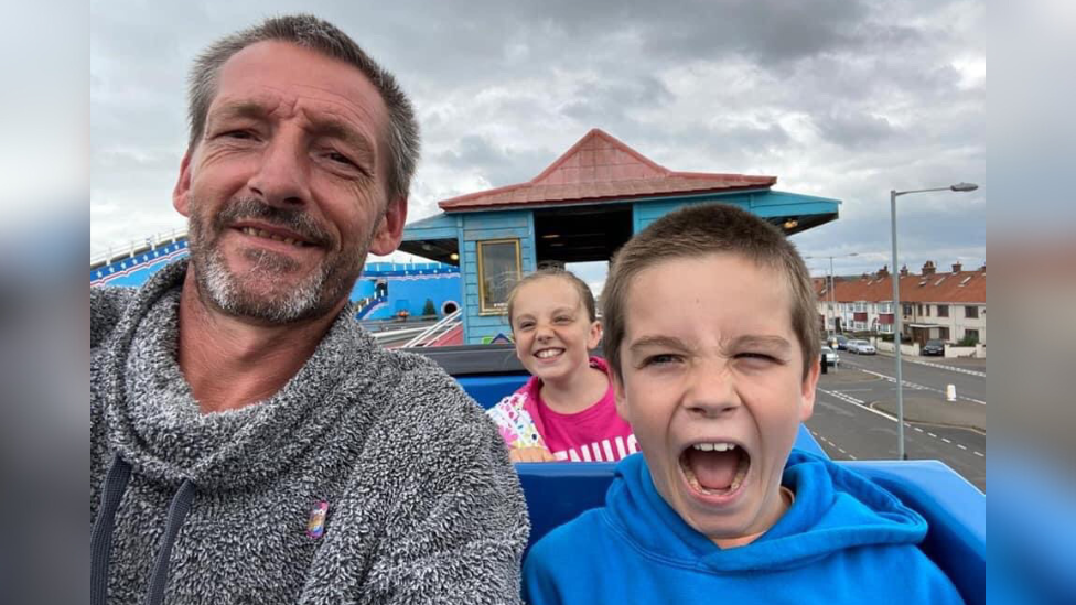 Jason Coleman, Freddie Coleman and a young girl sat on what appears to be a rollercoaster at a theme park. Jason is wearing a grey and white hoody, has short grey hair and is smiling. Freddie is younger than 15 years old here. He has short brown hair, is wearing a blue hoodie and has his mouth open in excitement. The young girl is smiling while wearing a pink T-shirt. She has light coloured hair which has partially blown across her face.