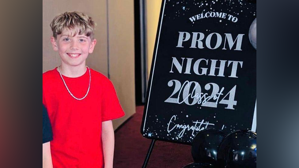 Mylo in a red t-shirt and chain, smiling at the camera, next to a big black sparkly board which says 'Welcome to Prom Night Class of 2024, Congratulations!'