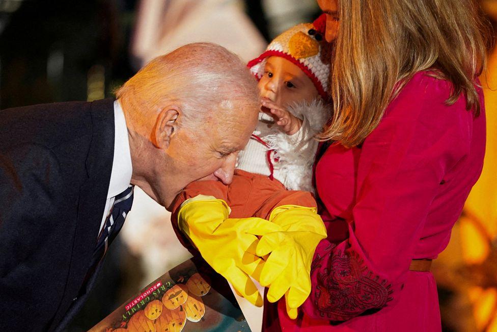 US President Joe Biden, wearing a dark suit, leans down to playfully bite a baby's leg. The baby is dressed in a chicken costume and is being held by a woman with long brown hair wearing a maroon-coloured dress