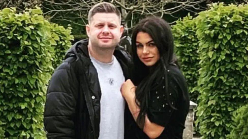 Josh and Chloe Bashford posing for photo in coats in front of bushes